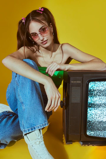 Tendência Y2K: a nostalgia dos anos 2000. Na foto, uma jovem usando top tank e calça reta, penteado com bumps e apoiada na tv de tubo. Reprodução/FreePik.